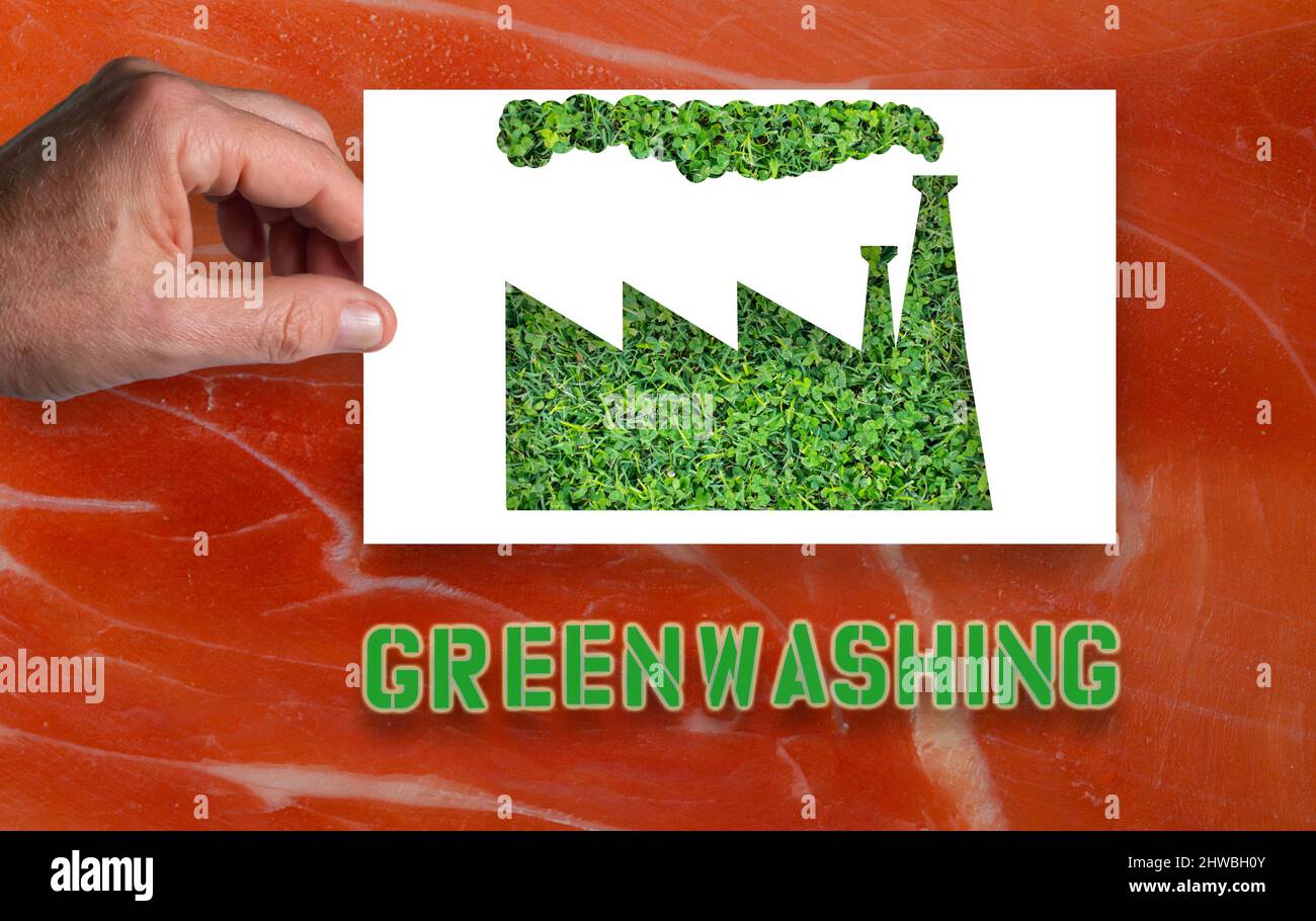 Greenwashing:`s Hände eines Mannes hält ein weißes Blatt Papier mit dem Umriss einer Fabrik, die innen mit grünem Gras ausgeschnitten ist.Hintergrund mit Schinkenfleisch Stockfoto